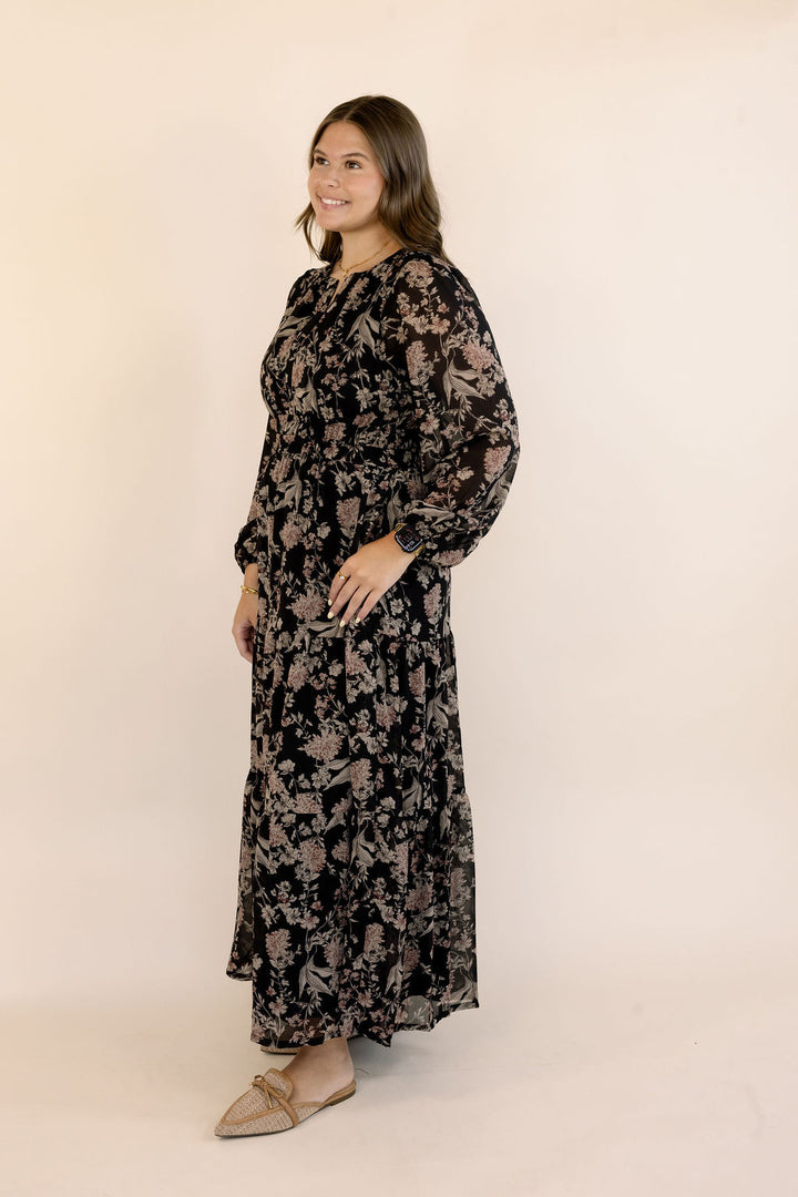 A woman modeling a long-sleeved black maxi dress with a muted floral pattern, paired with beige pointed flats, set against a plain beige studio background.