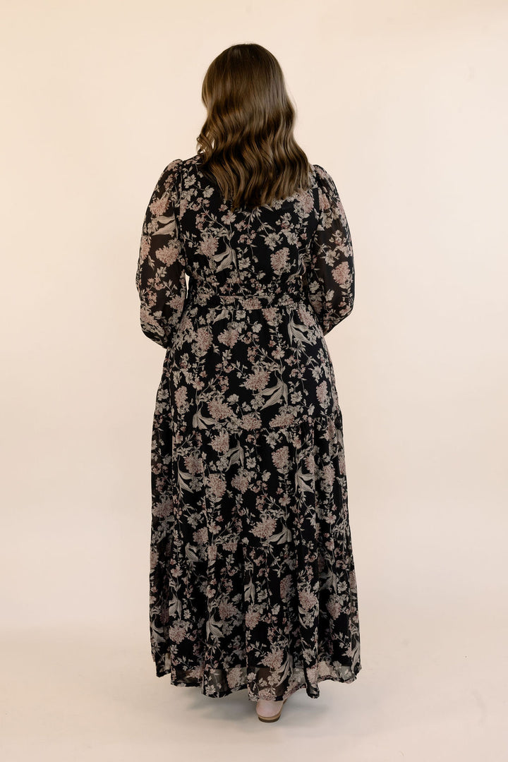 A woman viewed from behind wearing a long-sleeved black maxi dress with a beige floral pattern, standing against a plain beige studio background.