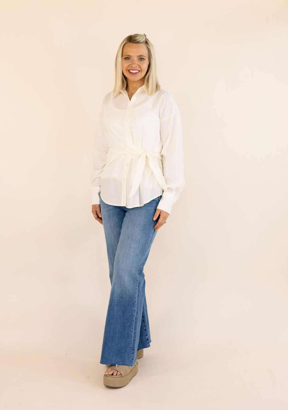 A woman modeling a cream long-sleeve button-up shirt with a tie waist, paired with blue flared jeans and beige platform sandals, against a solid light beige background.