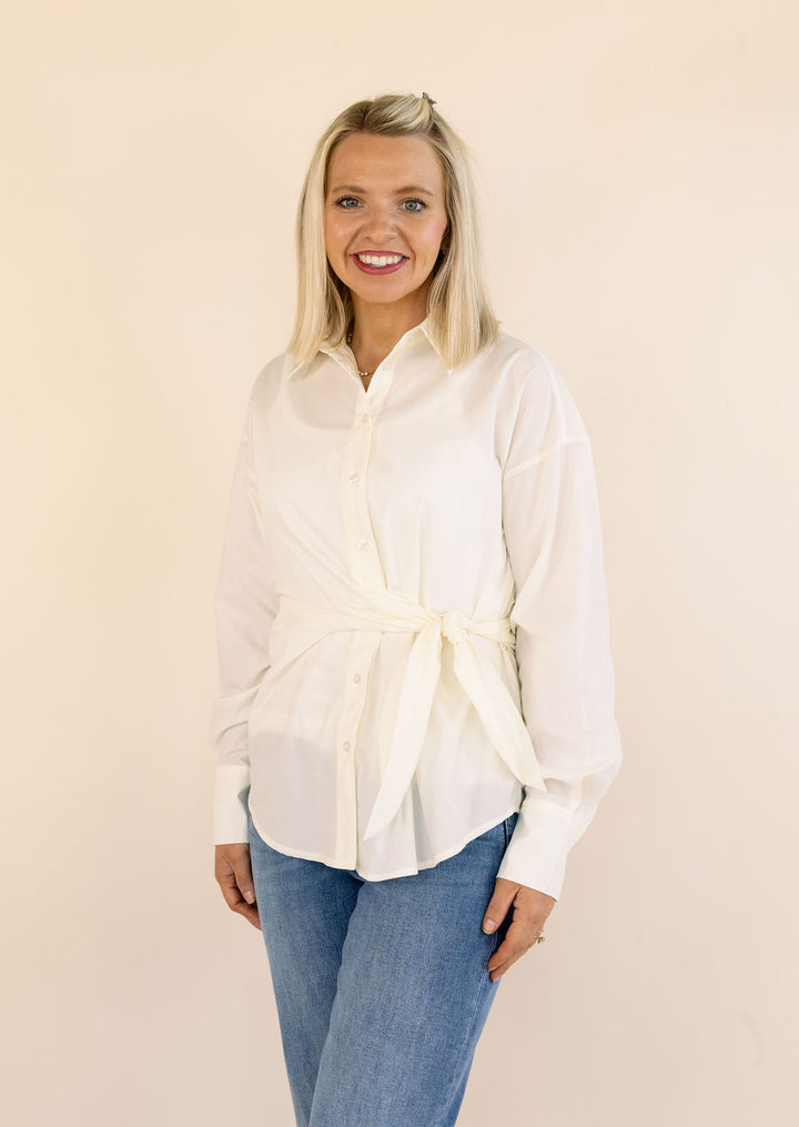 A smiling blonde woman wearing a cream long-sleeve button-up shirt with a tie waist and blue jeans, standing against a plain beige studio background.