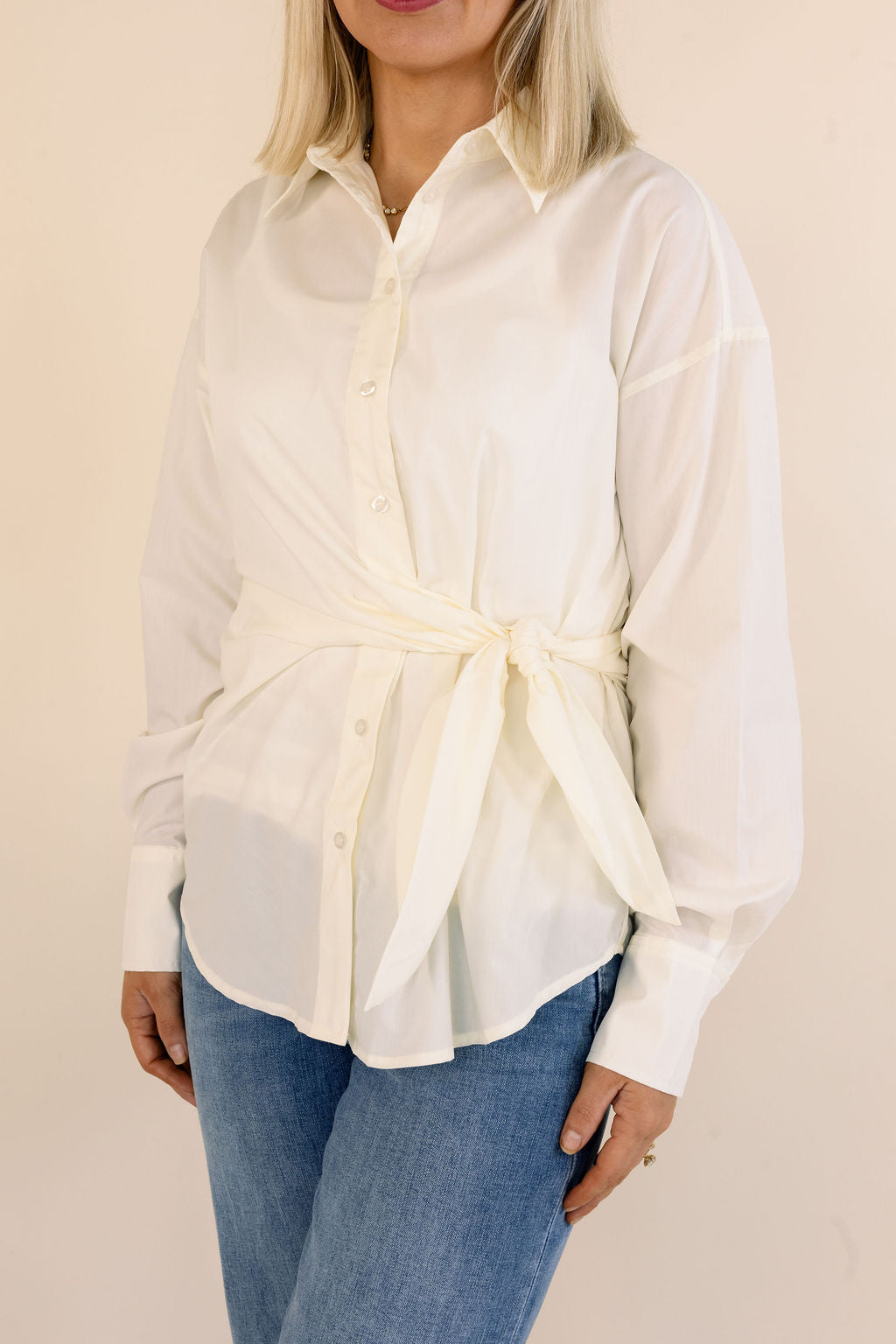 A woman wearing a cream-colored long-sleeve button-up shirt with a tie waist detail, paired with blue jeans, against a solid light beige background.