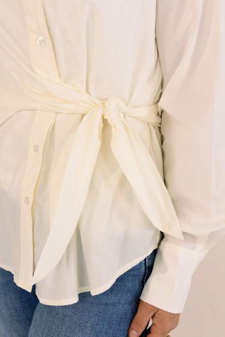 A close-up of a person wearing a cream-colored long-sleeve button-up shirt with a tie at the waist, paired with blue jeans.
