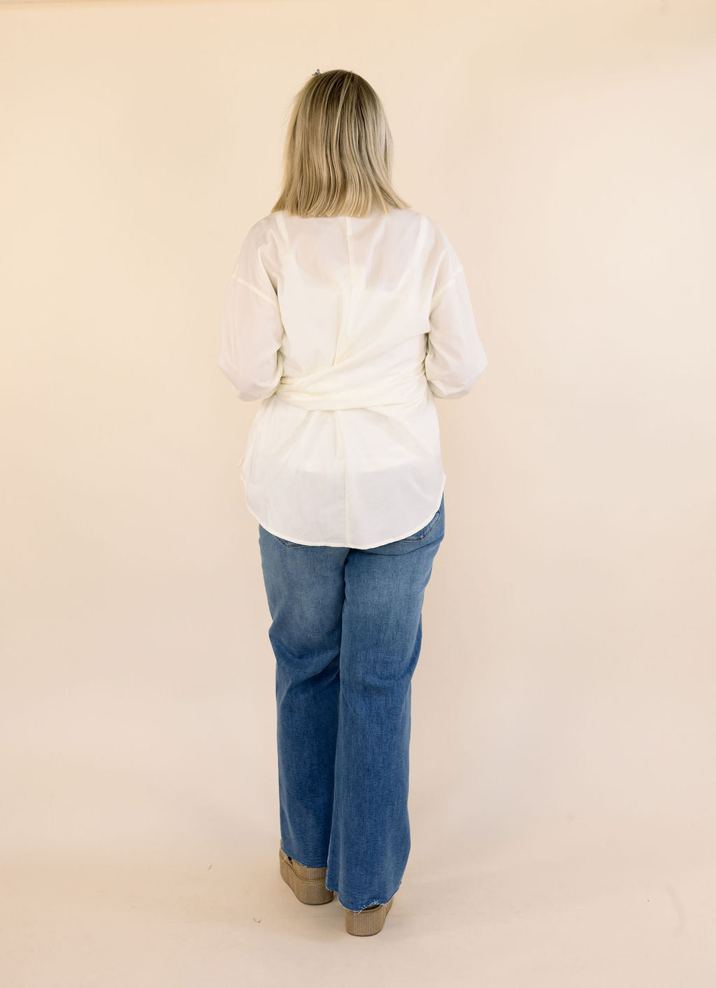 A woman viewed from behind wearing a cream long-sleeve side-tie blouse and blue jeans, standing against a plain beige studio background.