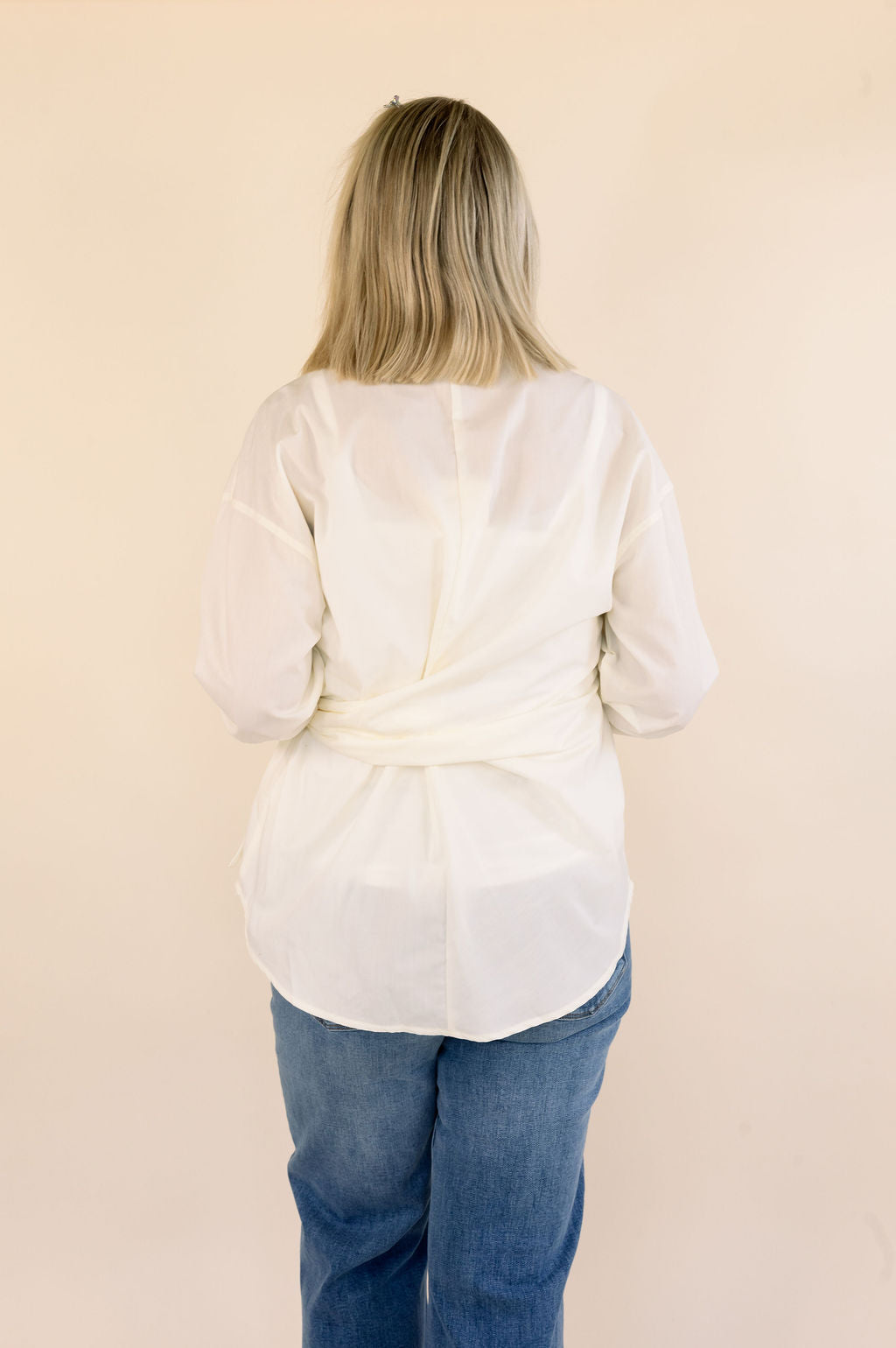 A woman viewed from behind wearing a cream long-sleeve side-tie blouse and blue jeans, standing against a plain beige background.