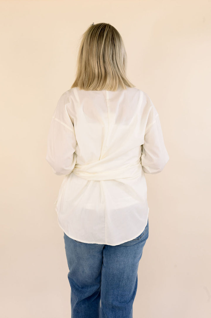 A woman viewed from behind wearing a cream long-sleeve side-tie blouse and blue jeans, standing against a plain beige background.