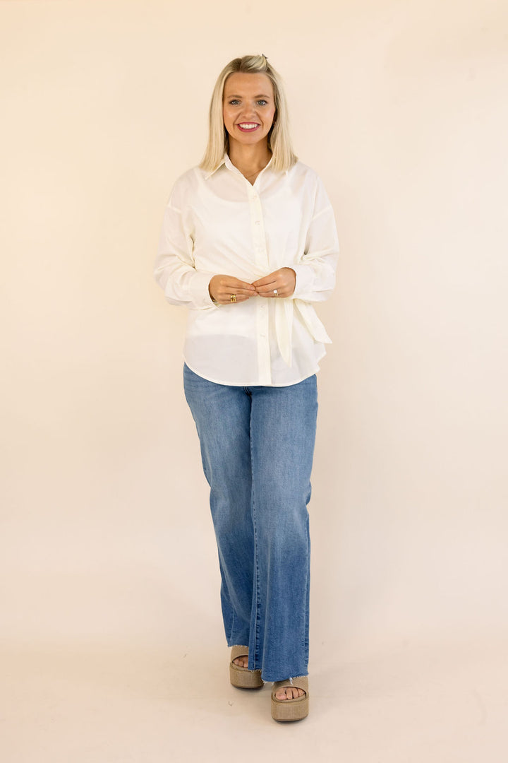 A woman modeling a cream long-sleeve button-up shirt and blue jeans against a solid beige studio background.