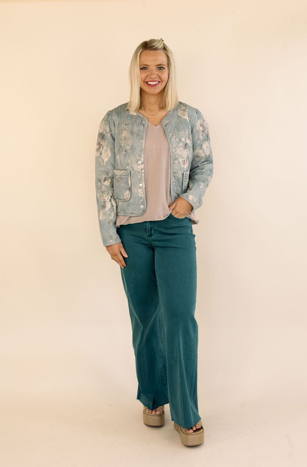 A woman modeling a light blue and white floral patterned bomber jacket, paired with a dusty pink top and green wide-leg jeans, standing against a solid cream-colored studio background.