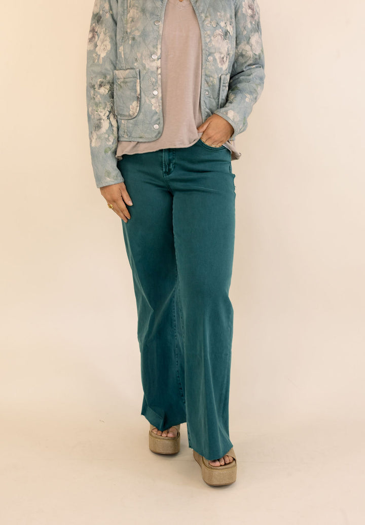 A model is shown from the chest down wearing dark green wide-leg jeans, a dusty pink top, a light blue floral jacket, and beige platform sandals, set against a plain beige studio background.