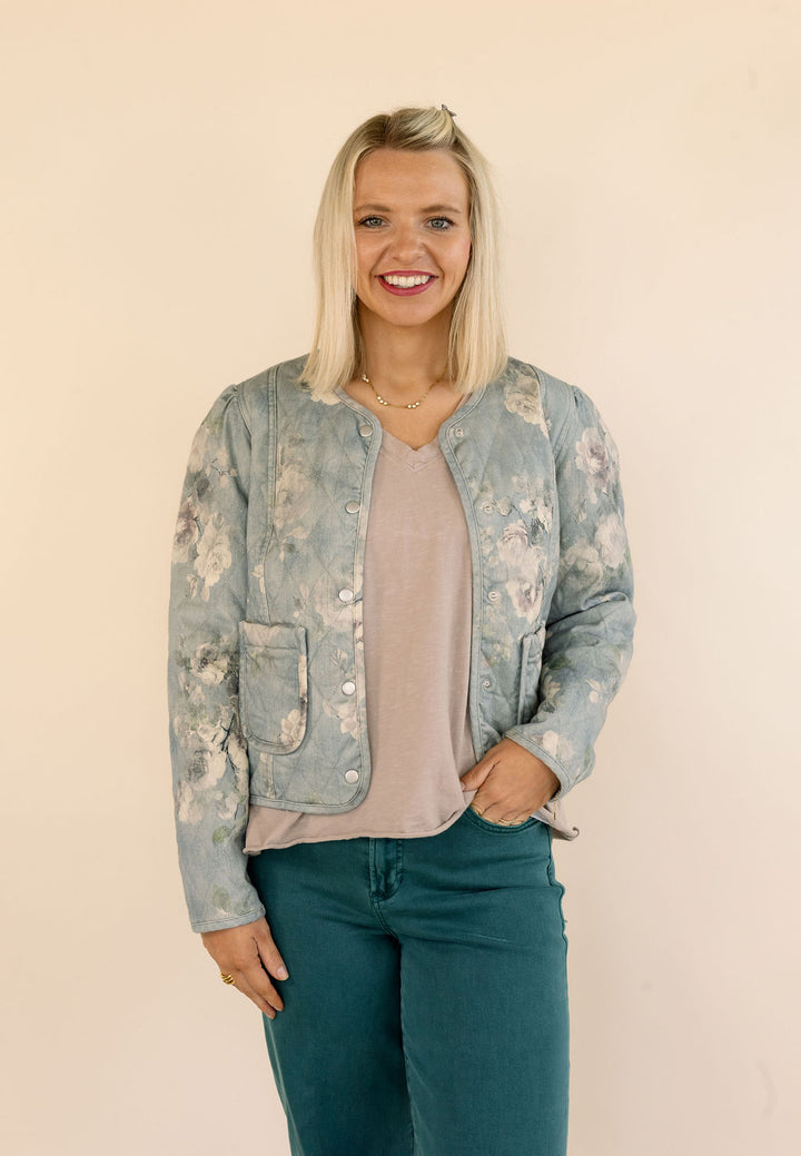 A smiling blonde woman wearing a light blue floral bomber jacket over a dusty pink top and green jeans, standing against a plain beige background.