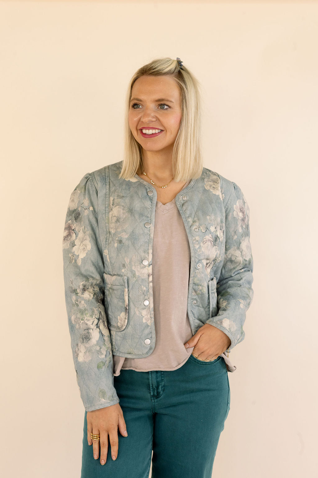 A woman with blonde hair wearing a light blue floral bomber jacket, a dusty pink top, and green jeans, standing against a solid beige background.