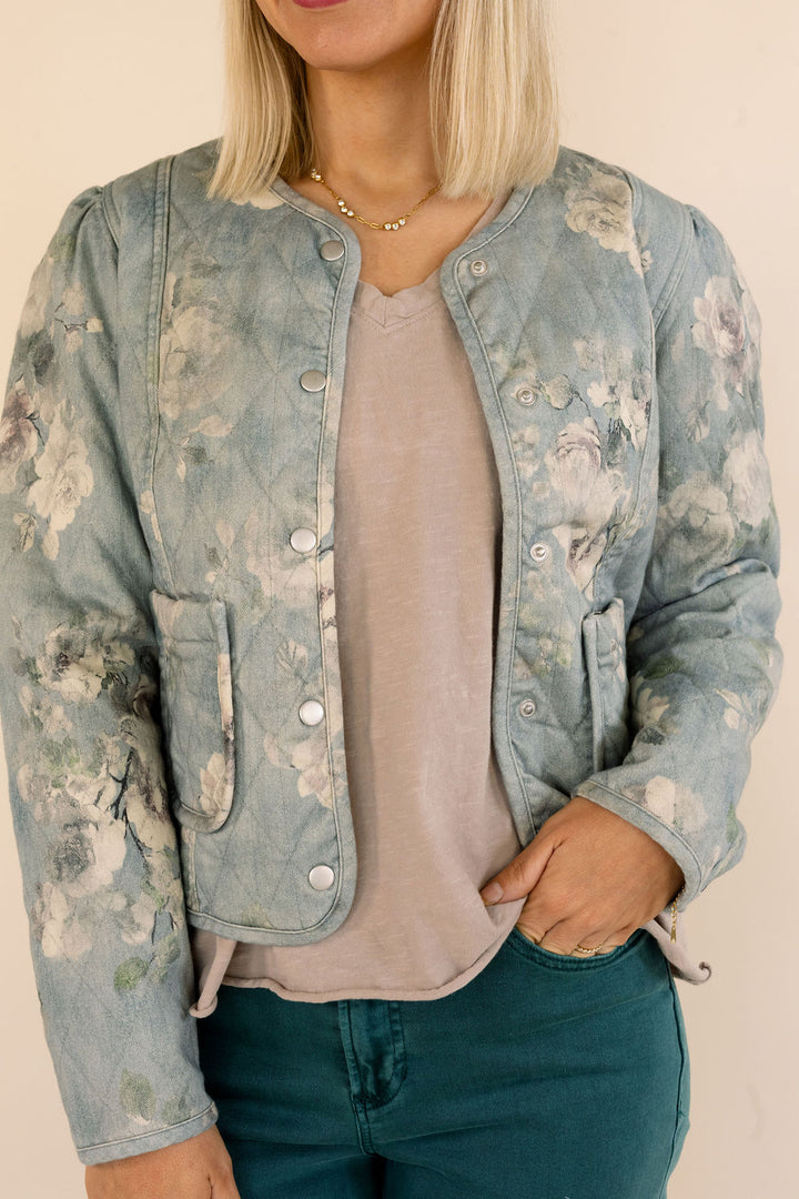 A woman wearing a light blue floral denim jacket over a dusty pink top and green jeans, shown from the neck down against a plain beige background.