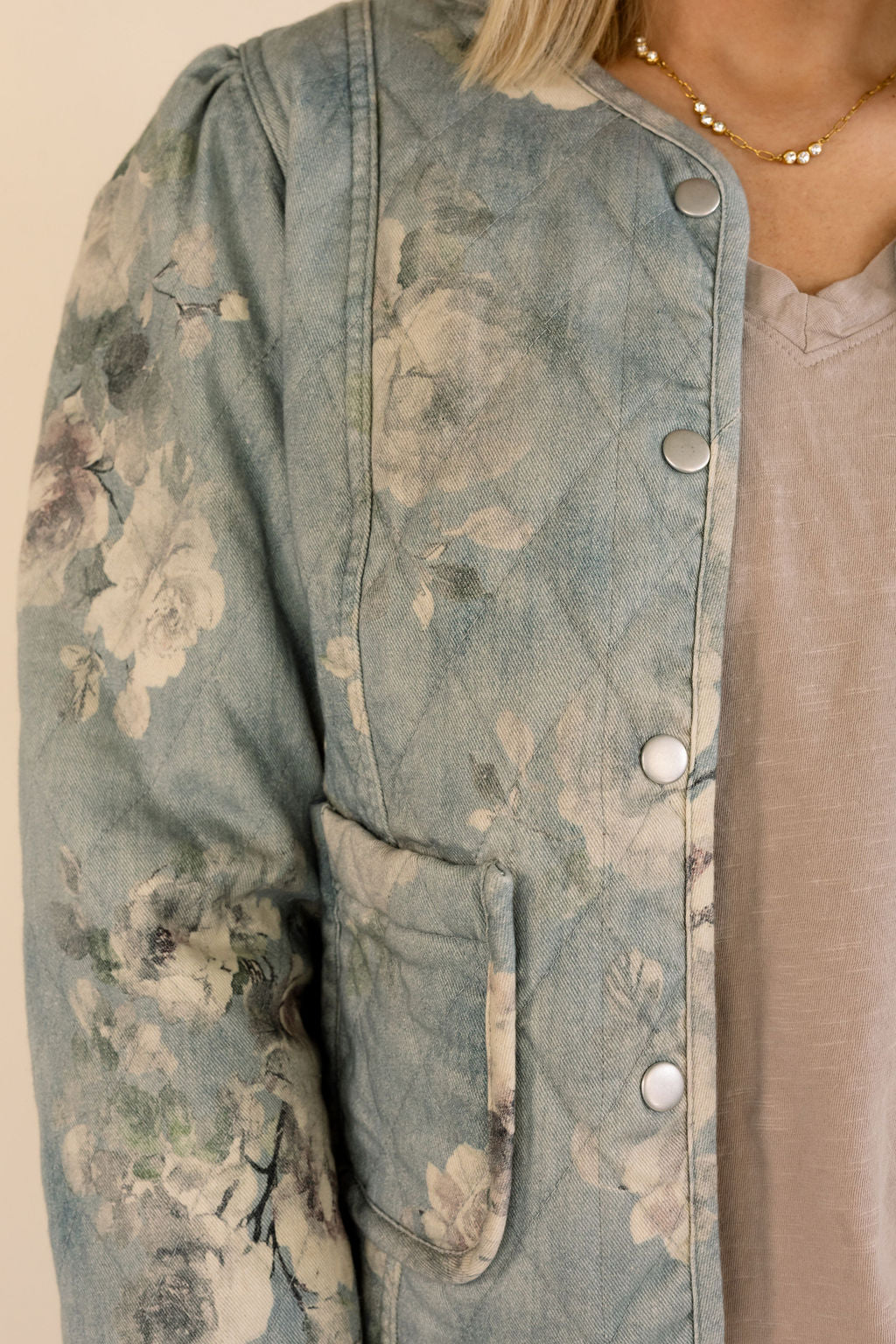 A close-up of a person wearing a light blue floral quilted jacket over a taupe top, with a gold beaded necklace visible.