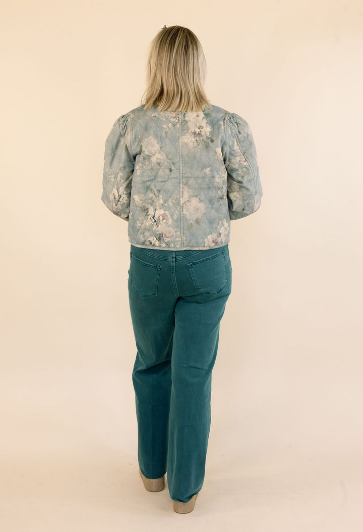 A woman viewed from behind wearing a light blue floral print jacket with puffed sleeves and green jeans, standing against a plain beige studio background.