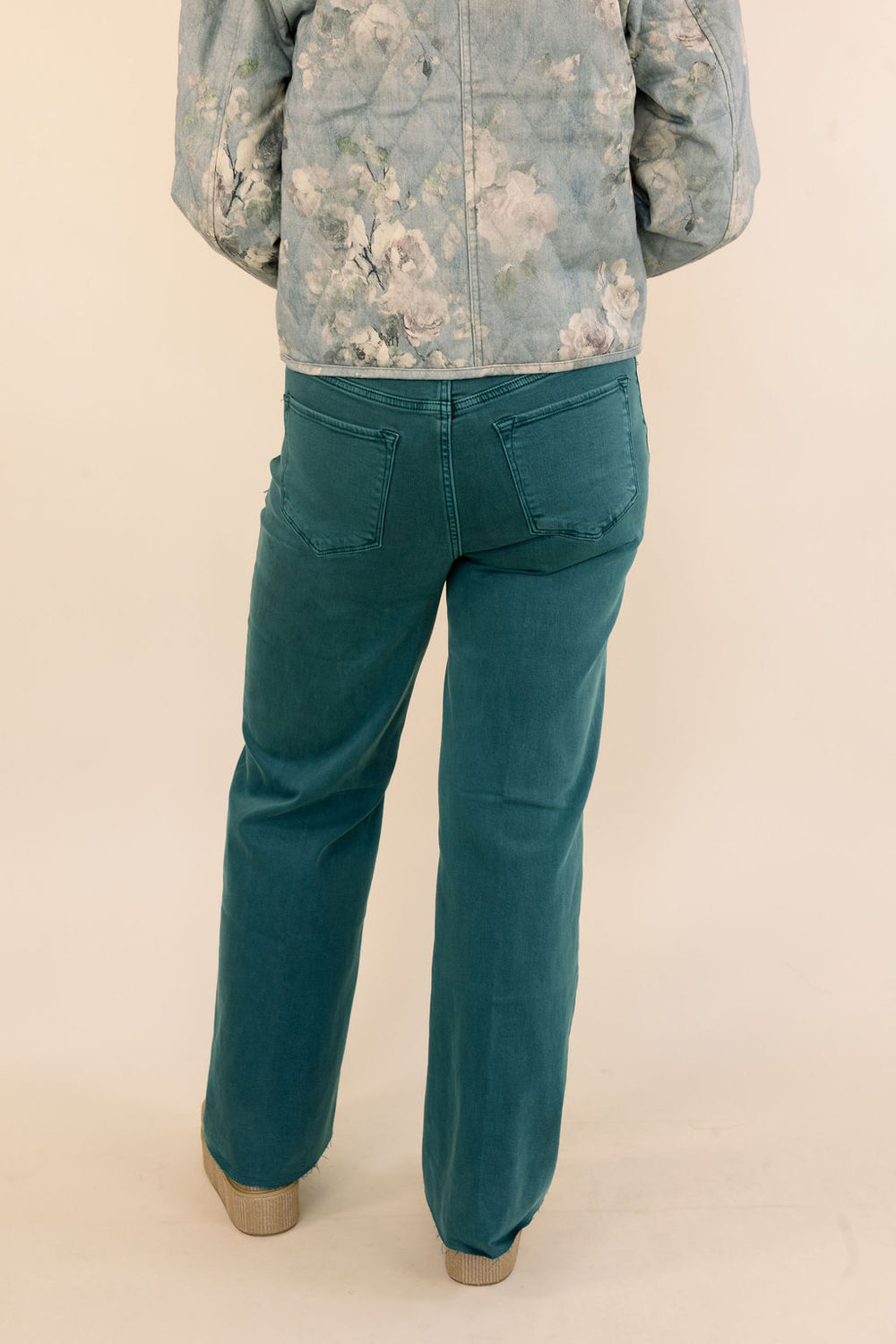 A person shown from the back wearing dark green wide-leg jeans and a light blue floral jacket, standing against a plain beige studio background.