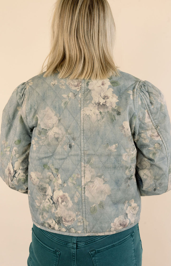 A person shown from the back wearing a light blue floral quilted jacket with puff sleeves and green jeans, set against a plain beige background.
