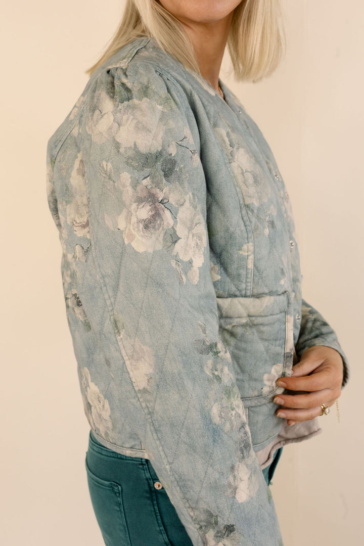 A woman wearing a light blue floral quilted jacket with a faded, washed texture, paired with green jeans, shown from a side profile against a plain beige background.