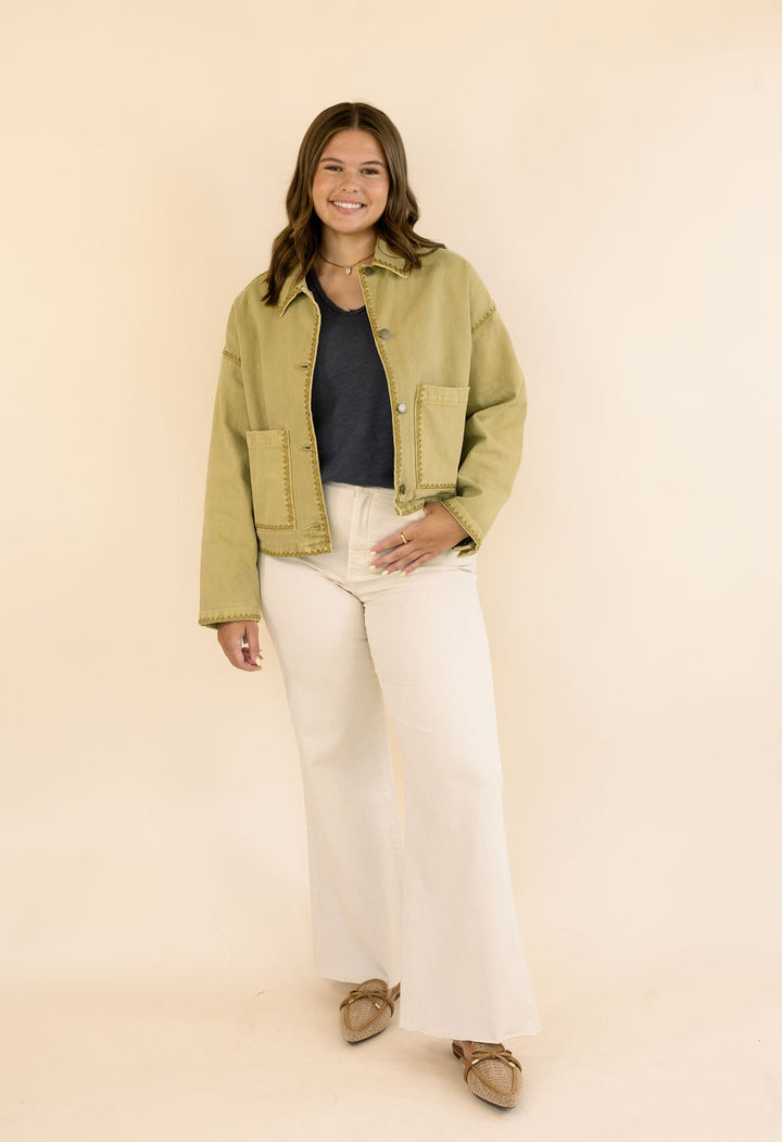 A woman modeling a khaki cropped utility jacket over a dark navy top and cream wide-leg jeans, standing against a solid warm beige studio background.