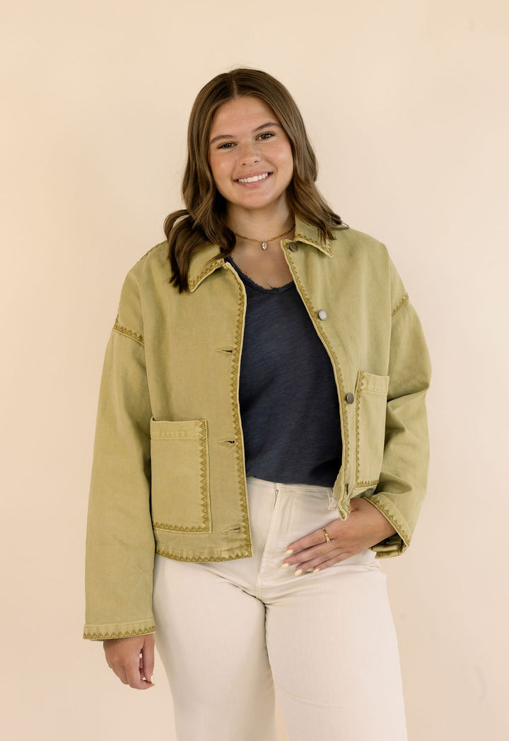 A woman modeling a cropped, khaki denim jacket with decorative gold-toned stitching, worn over a dark blue top and cream-colored jeans, set against a solid beige studio background.