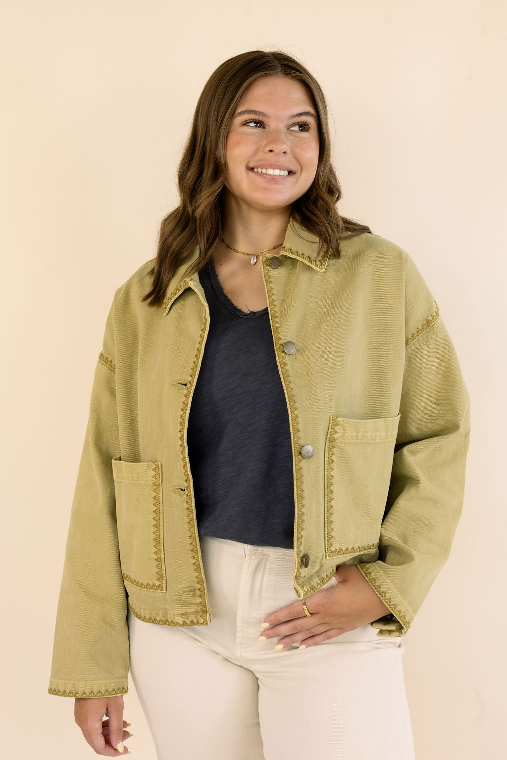 A woman wearing a cropped khaki denim jacket with decorative stitching, a dark gray top, and cream jeans, standing against a solid beige background.