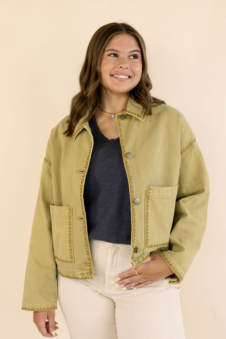 A woman wearing a cropped khaki denim jacket with decorative stitching, a dark gray top, and cream jeans, standing against a solid beige background.