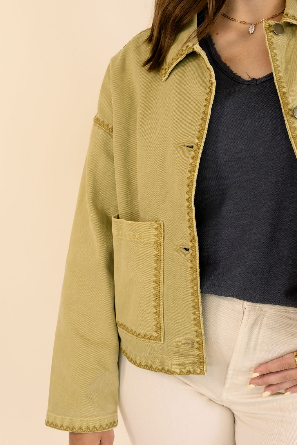 A cropped view of a person wearing a khaki jacket with decorative zigzag stitching, paired with a dark blue top and cream pants, against a solid beige background.