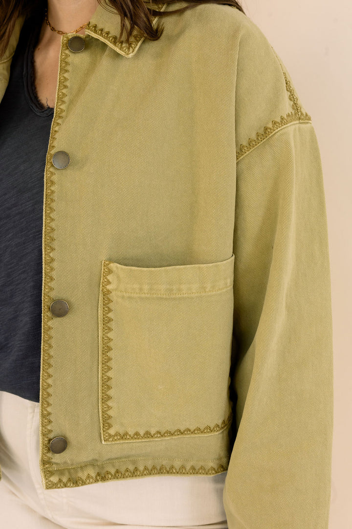 A cropped khaki jacket with decorative zigzag stitching and dark buttons, worn by a model over a dark top and white pants.