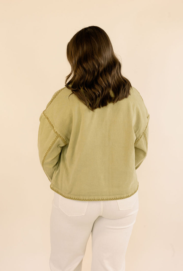 A woman viewed from behind wearing a khaki cropped jacket with decorative stitching and white pants, set against a plain beige background.