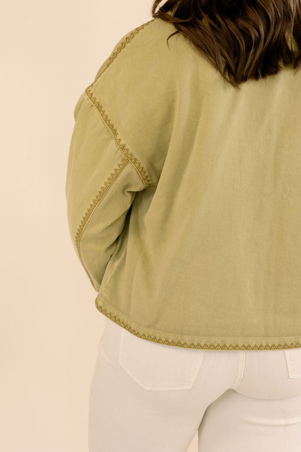 Close-up back view of a person wearing a cropped khaki jacket with decorative gold-colored stitching and white pants, against a plain beige background.