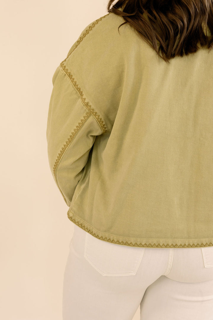 Close-up back view of a person wearing a cropped khaki jacket with decorative gold-colored stitching and white pants, against a plain beige background.