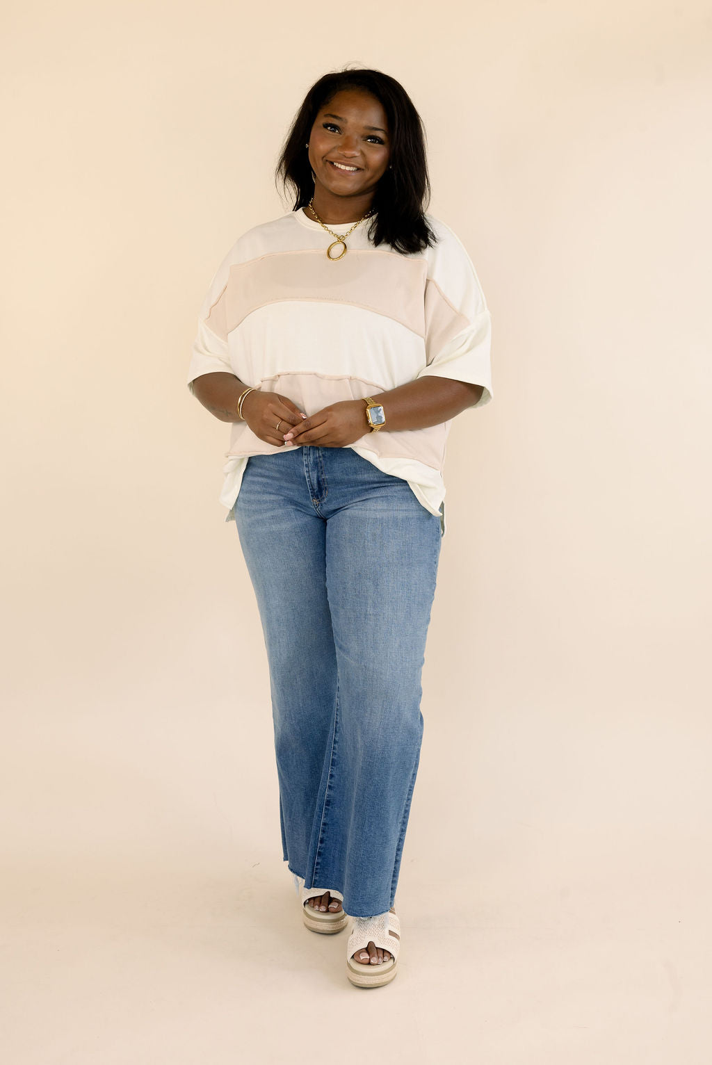 Front view of plus size short sleeve color block top in taupe and cream, styled with blue denim jeans and gold accessories.