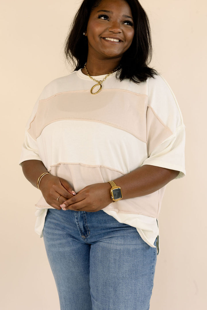 Front view of plus size taupe and cream color block top with round neckline, dropped shoulders, and short sleeves, paired with denim jeans.