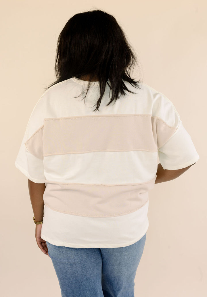 Back view of plus size taupe and cream color block short sleeve top featuring horizontal panels and exposed seams, styled with blue jeans.
