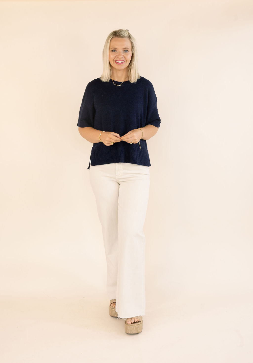 A woman modeling a short-sleeve navy blue knit top and off-white wide-leg pants against a solid light beige studio background.