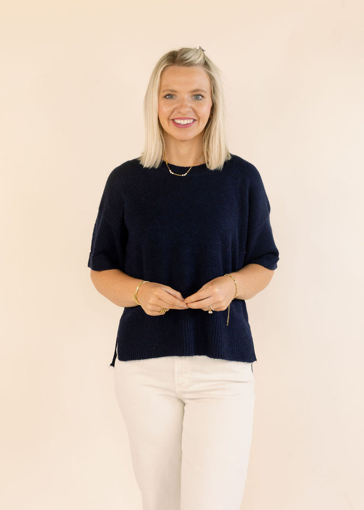 A smiling blonde woman wearing a navy blue short-sleeve knit top and cream pants, standing against a plain beige background.
