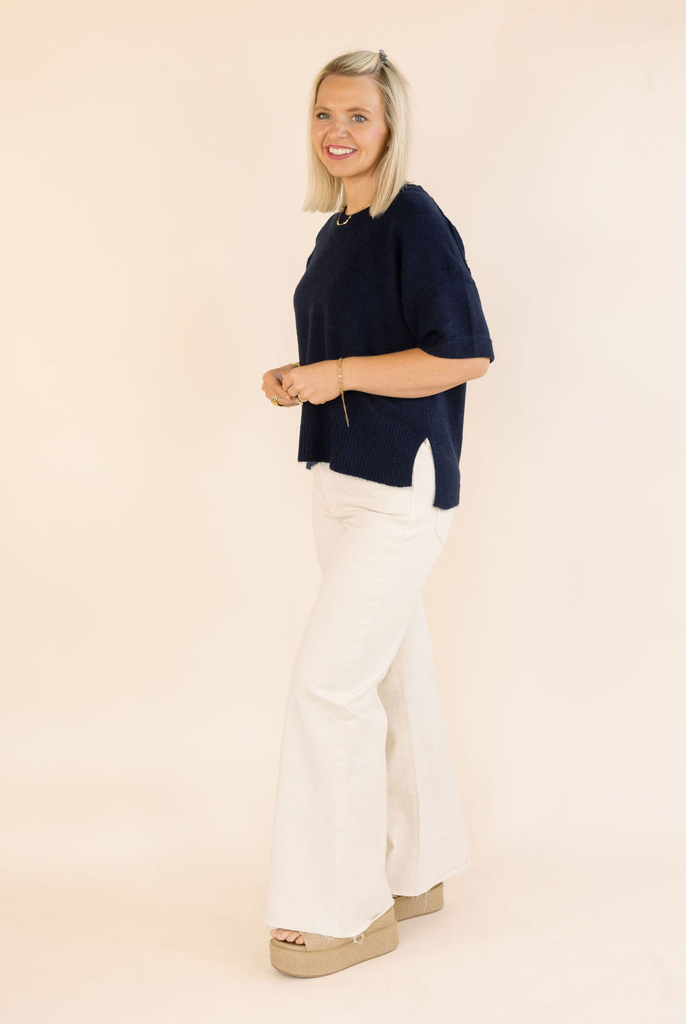 A woman modeling a navy blue short-sleeve knit top and off-white wide-leg jeans against a solid light beige studio background.