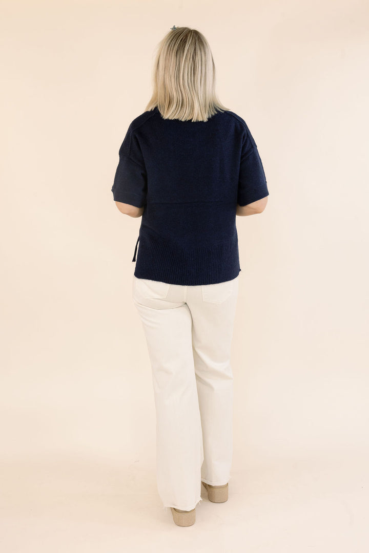 A woman viewed from behind wearing a short-sleeve navy blue knit top and off-white pants, standing against a plain beige studio background.