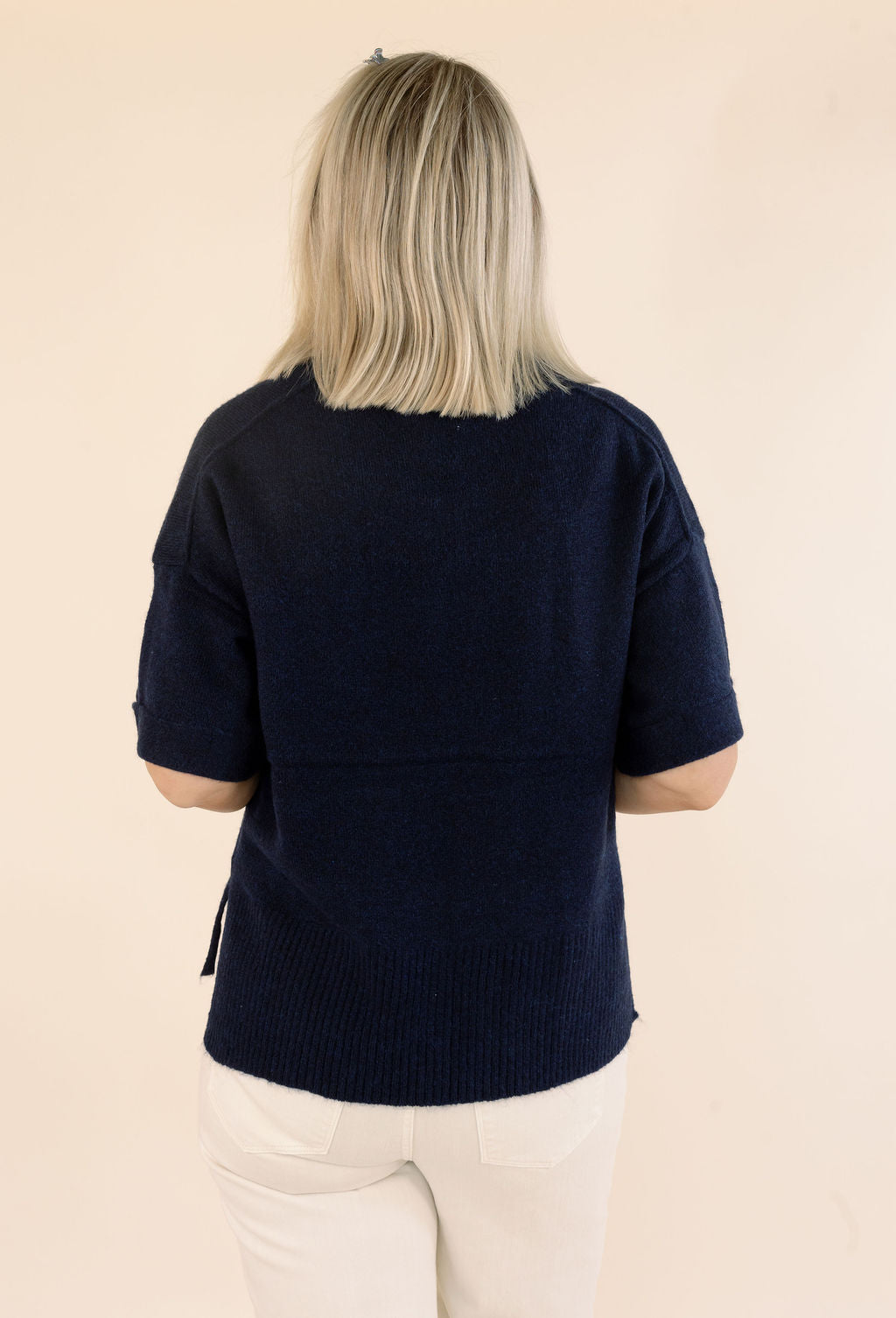A woman viewed from behind wearing a dark navy blue short-sleeve knit sweater and white pants, standing against a plain beige background.