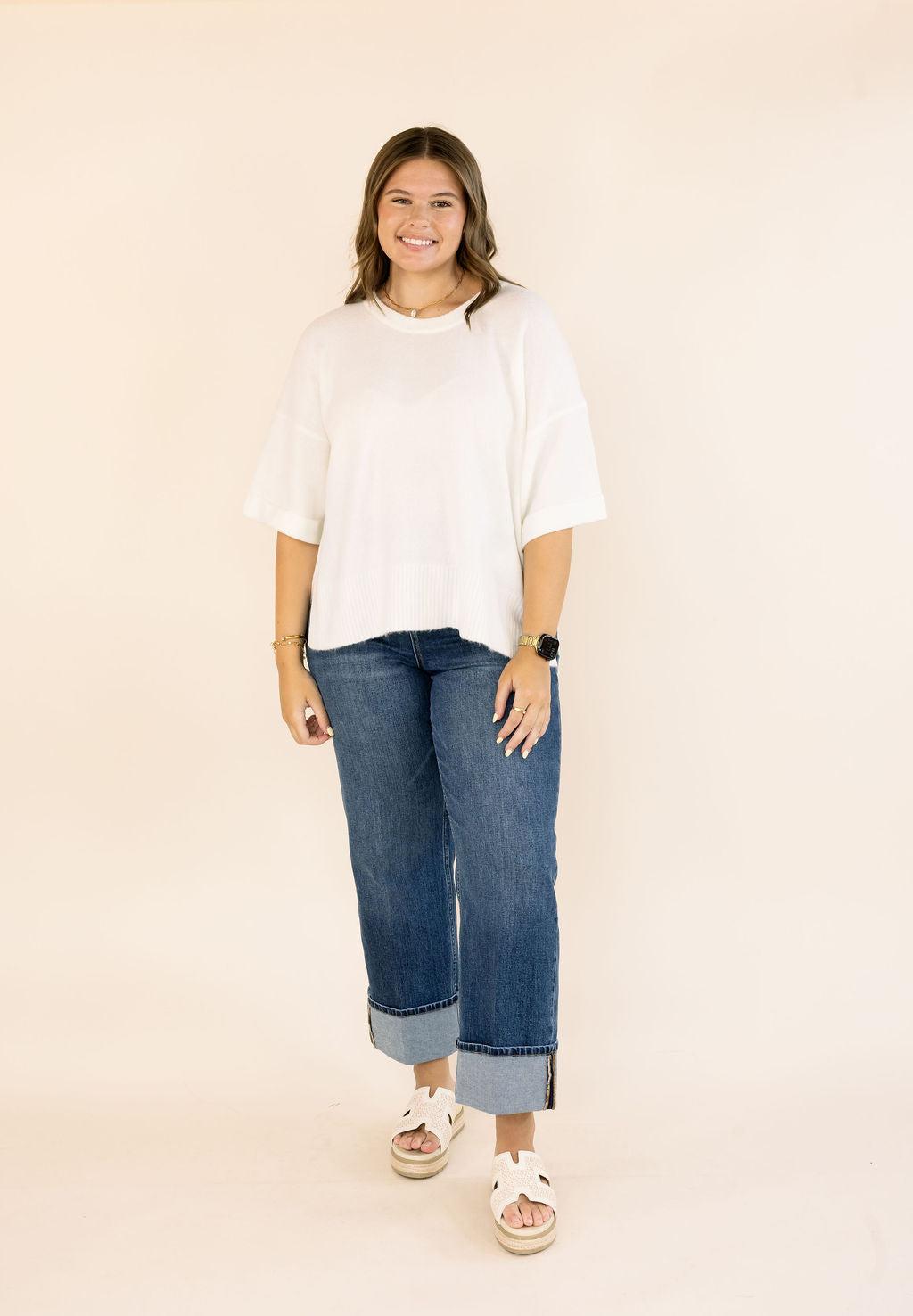 A woman modeling a n off-white short-sleeve crewneck sweater and blue cuffed jeans, standing against a solid light beige background.