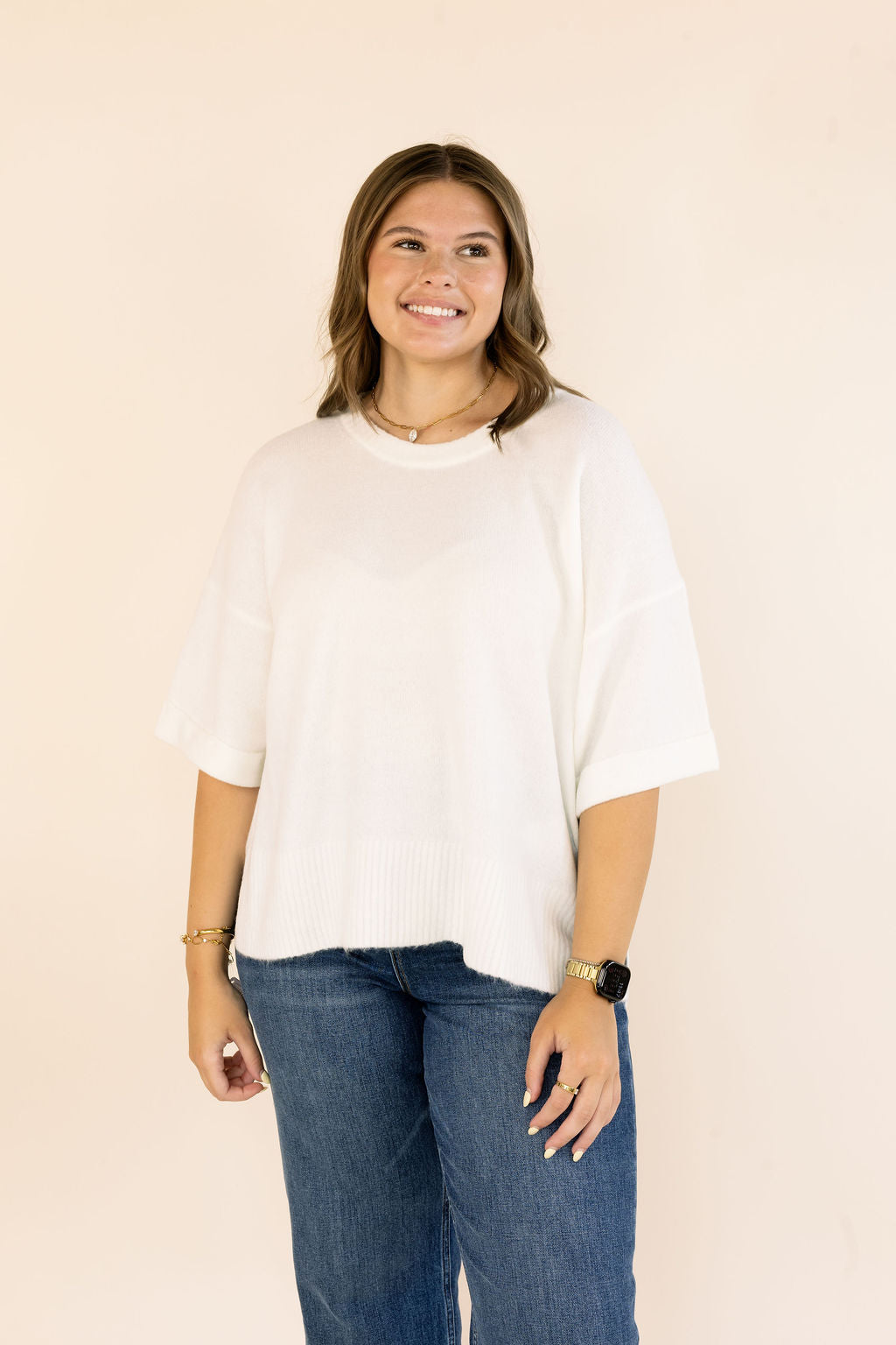 A woman wearing an off- white short-sleeve knit top and blue jeans, standing against a solid light beige background.