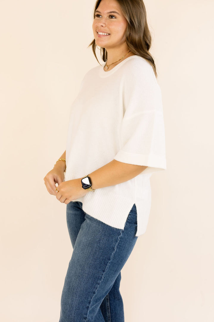 A woman wearing an off- white short-sleeve knit top with a side slit and blue jeans, standing against a plain light beige studio background.