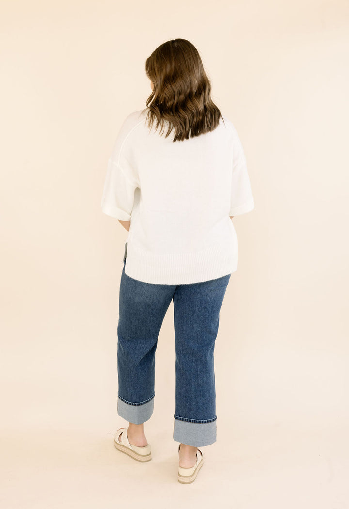A woman viewed from behind wearing an oversized off-white short-sleeve knit top, blue cuffed jeans, and white slip-on shoes against a plain beige studio background.