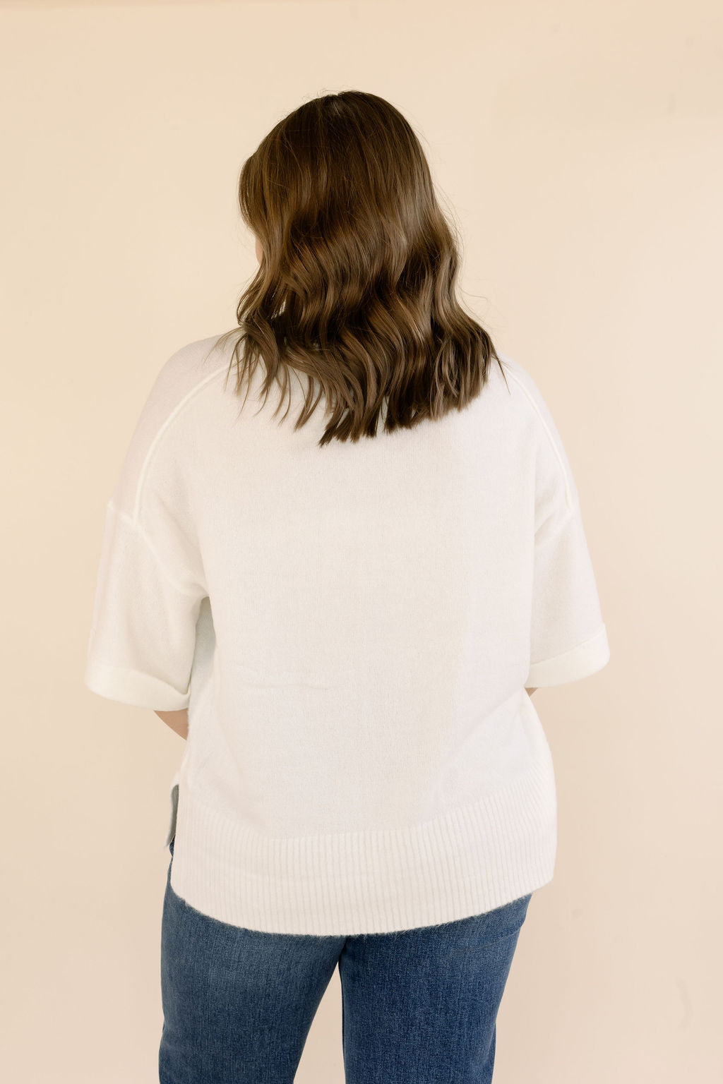 A woman viewed from behind wearing an off-white short-sleeve knit sweater and blue jeans against a plain beige background.