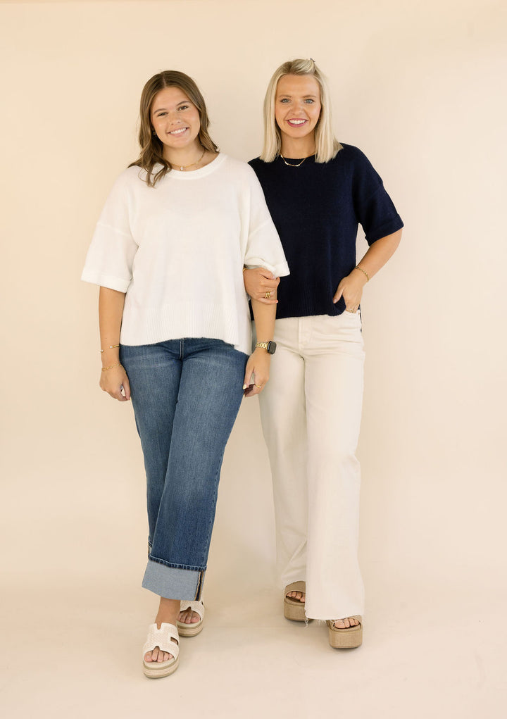 Two women standing side-by-side in a studio setting, wearing plain, oversized, short-sleeve sweaters in off-white and navy blue, paired with jeans and wide-leg trousers.