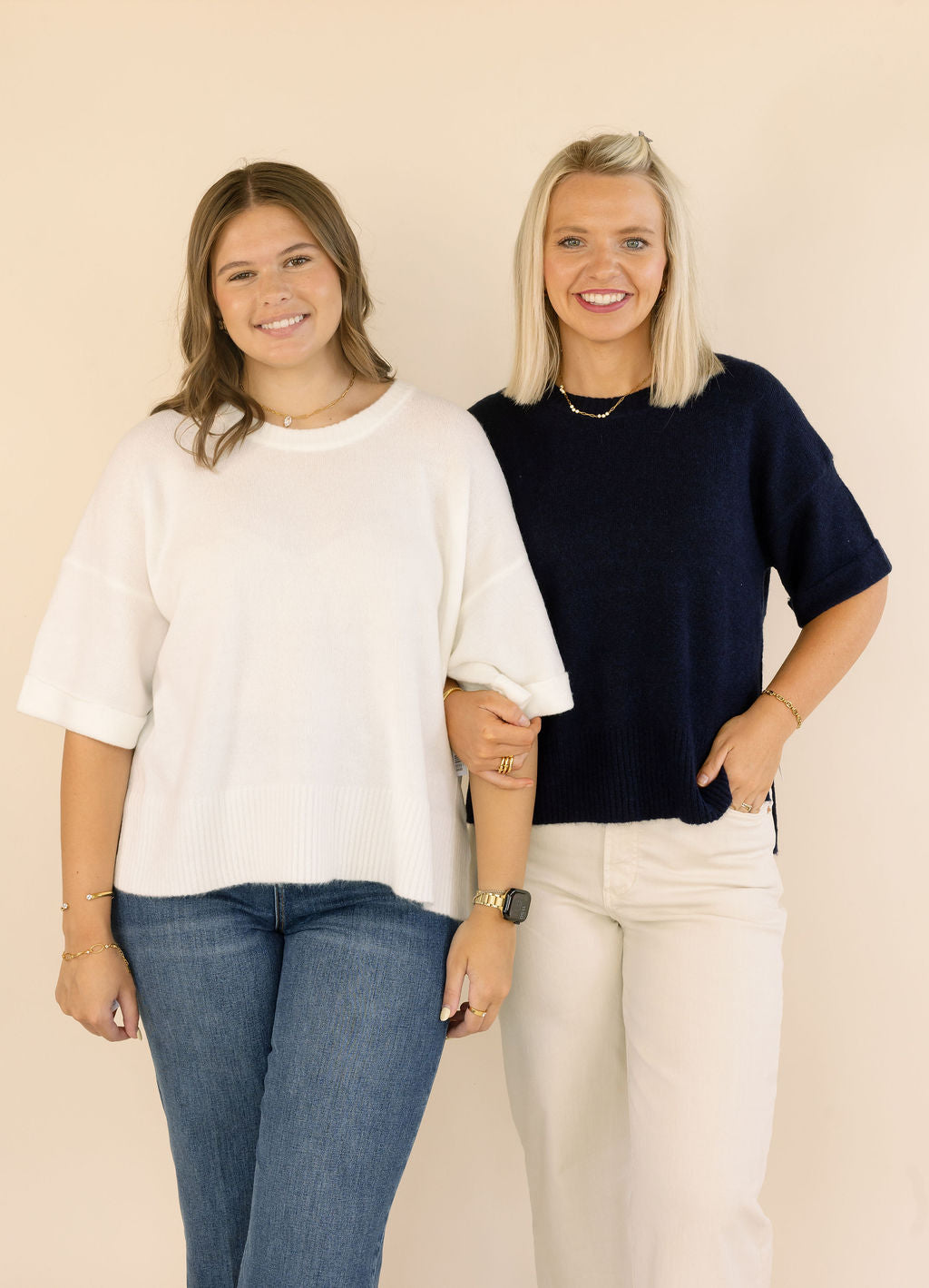 Two women standing side-by-side wearing knit crewneck sweaters, one in off-white and one in navy blue, paired with jeans and cream pants against a solid beige background.