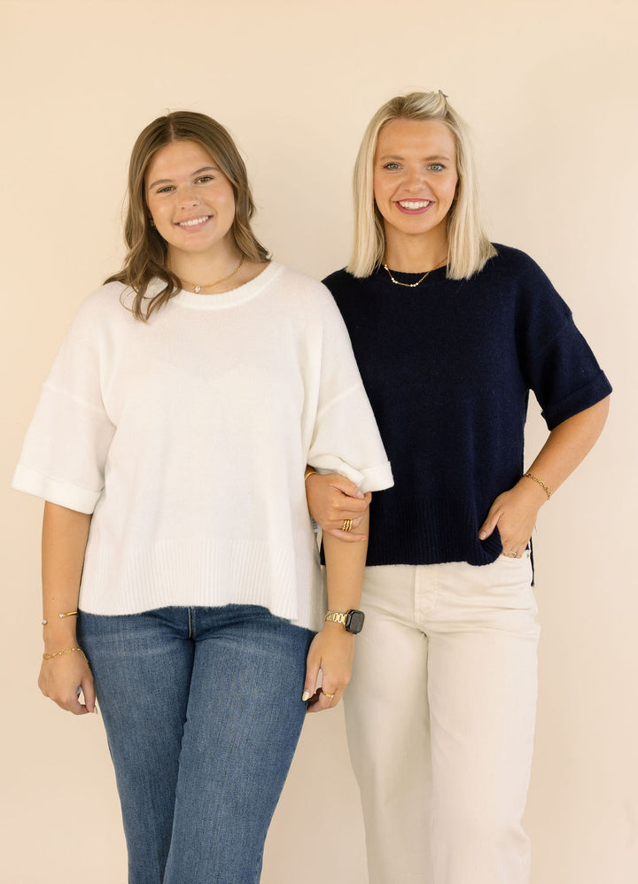 Two women standing side-by-side wearing knit crewneck sweaters, one in off-white and one in navy blue, paired with jeans and cream pants against a solid beige background.