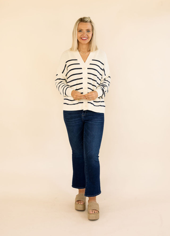 A woman modeling a cream cardigan sweater with navy blue horizontal stripes, paired with dark blue jeans and beige platform sandals, against a solid cream studio background.