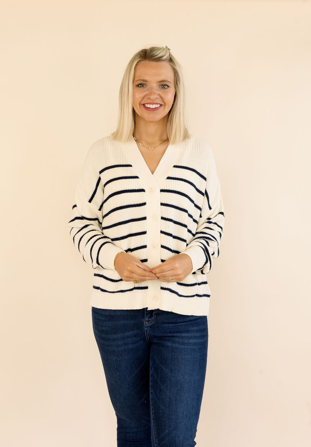 A smiling blonde woman wearing a cream-colored knit cardigan with navy blue horizontal stripes and dark blue jeans, standing against a solid light beige background.