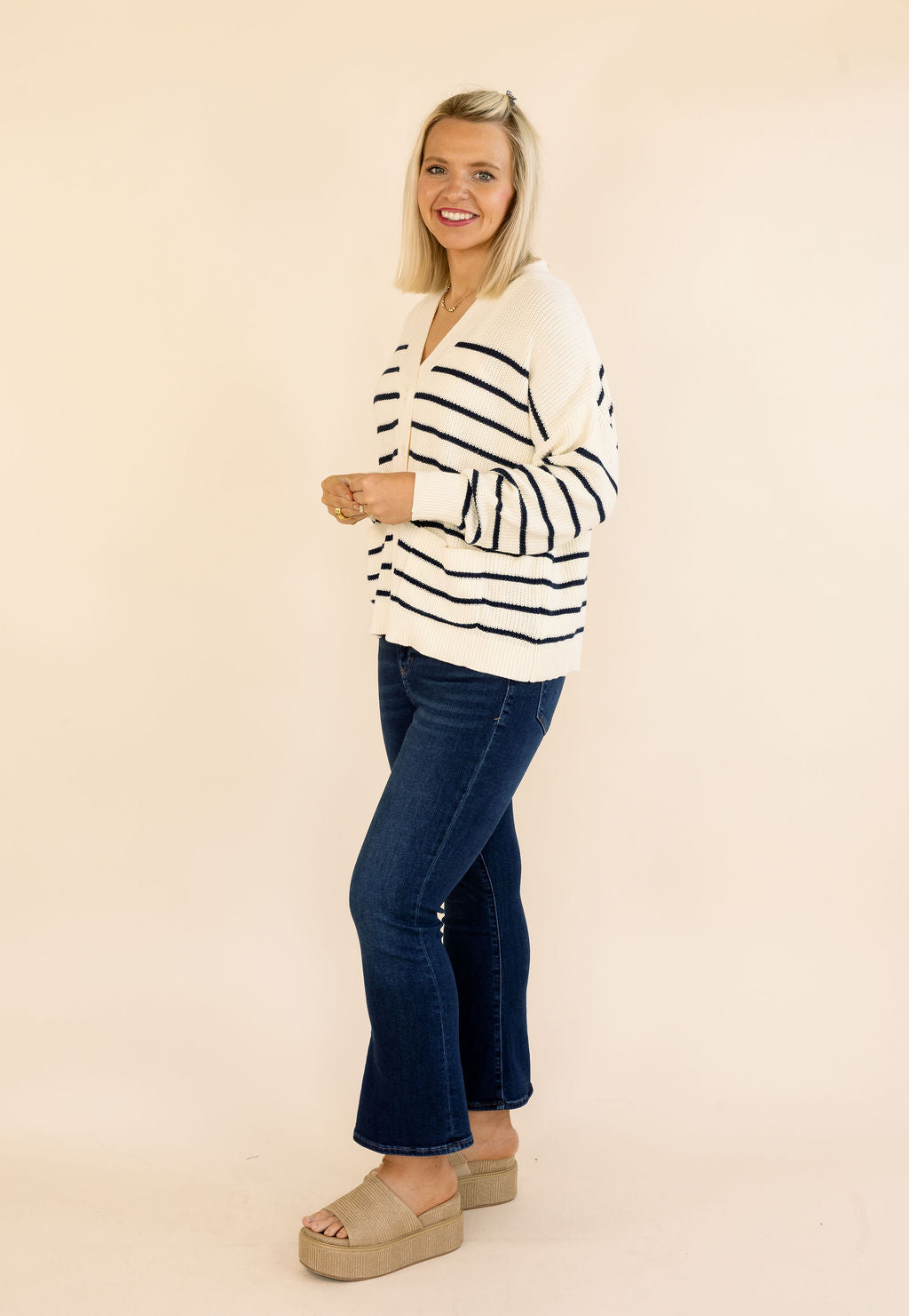A woman modeling a cream knit sweater with black horizontal stripes, paired with dark blue jeans and beige platform sandals, standing against a solid light beige background.