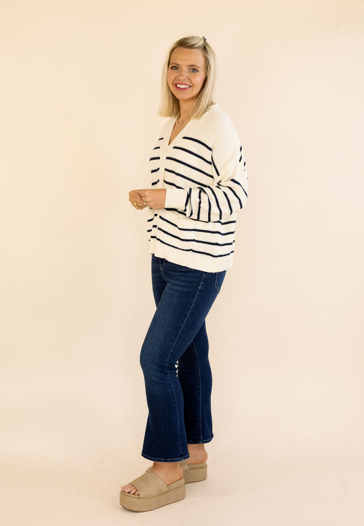 A woman modeling a cream knit sweater with black horizontal stripes, paired with dark blue jeans and beige platform sandals, standing against a solid light beige background.