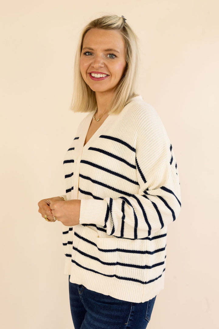 A smiling blonde woman wearing a cream-colored knit cardigan with navy blue horizontal stripes, paired with dark blue jeans, standing against a solid light beige background.